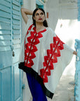 A person wearing a white kaftan with red geometric patterns, standing in front of a blue door.