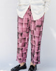 An older woman with short gray hair and glasses stands against a white studio background wearing a white button-up shirt, pink and black patterned trousers, and black loafers.