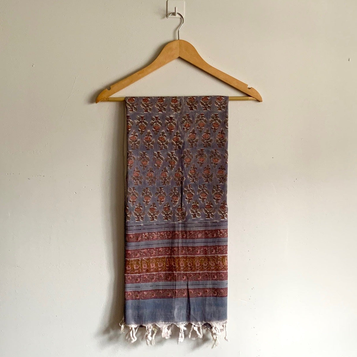A patterned scarf with blue, rust, and brown tones hanging from a wooden hanger against a plain off-white wall.