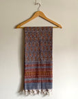 A patterned scarf with blue, rust, and brown tones hanging from a wooden hanger against a plain off-white wall.
