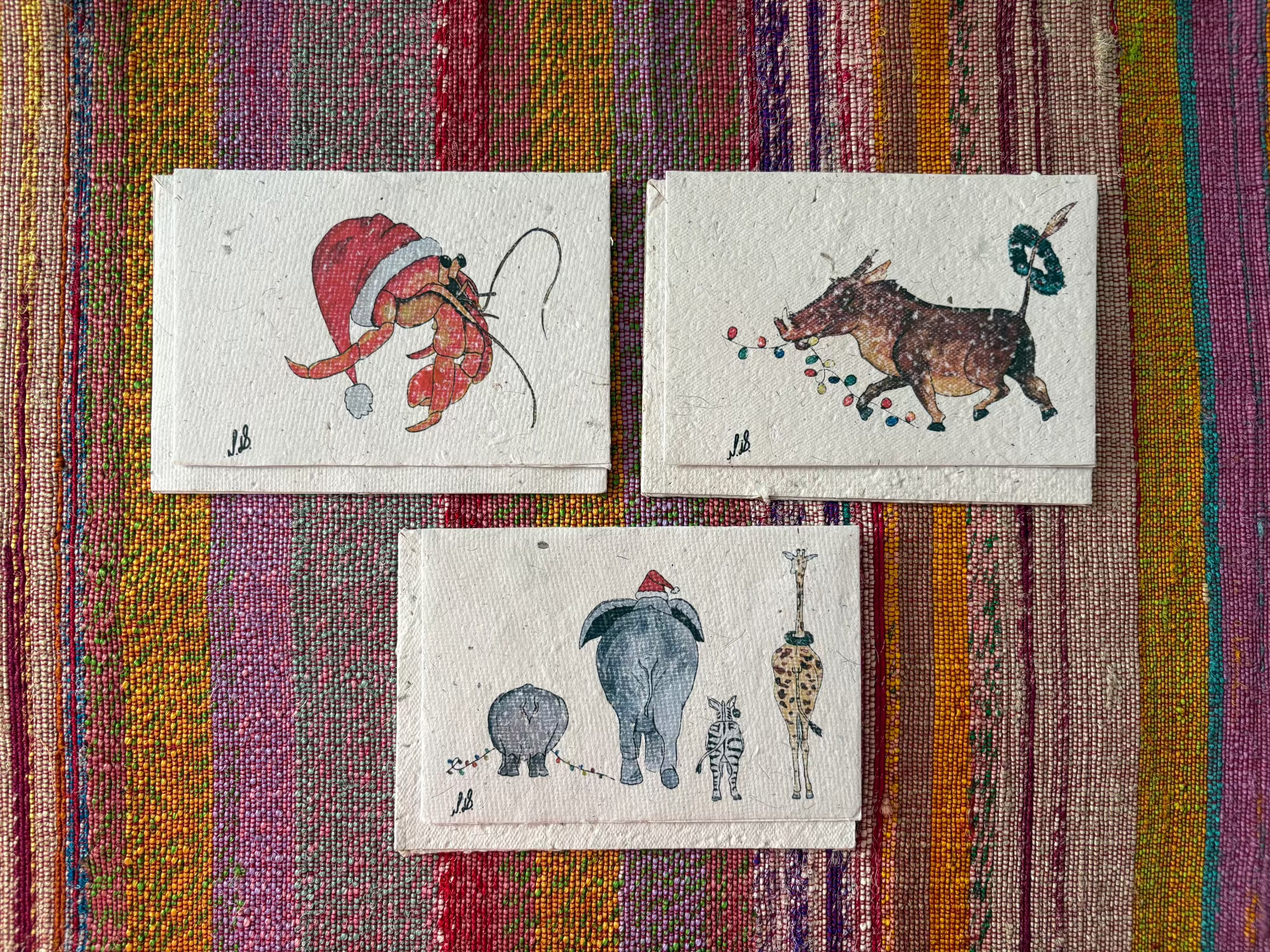 Greeting cards with illustrated animal drawings are arranged on a colorful striped woven textile background. The top-right card shows a red Santa hat and a hermit crab; the bottom-right card shows a warthog with colorful dots and a green wreath; the left card shows a rhino, elephant, zebra and giraffe.