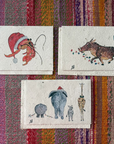 Greeting cards with illustrated animal drawings are arranged on a colorful striped woven textile background. The top-right card shows a red Santa hat and a hermit crab; the bottom-right card shows a warthog with colorful dots and a green wreath; the left card shows a rhino, elephant, zebra and giraffe.
