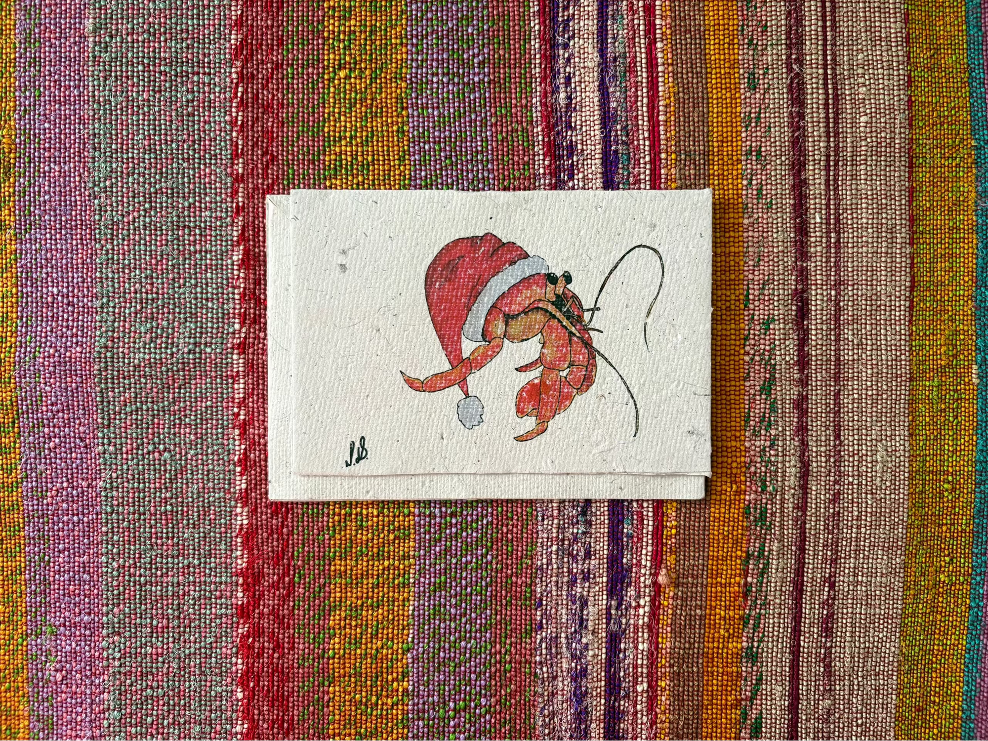A small handmade paper card featuring a whimsical illustration of a Santa Claus figure in a red Santa hat, placed on a colorful striped woven textile background.
