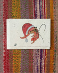 A small handmade paper card featuring a whimsical illustration of a Santa Claus figure in a red Santa hat, placed on a colorful striped woven textile background.