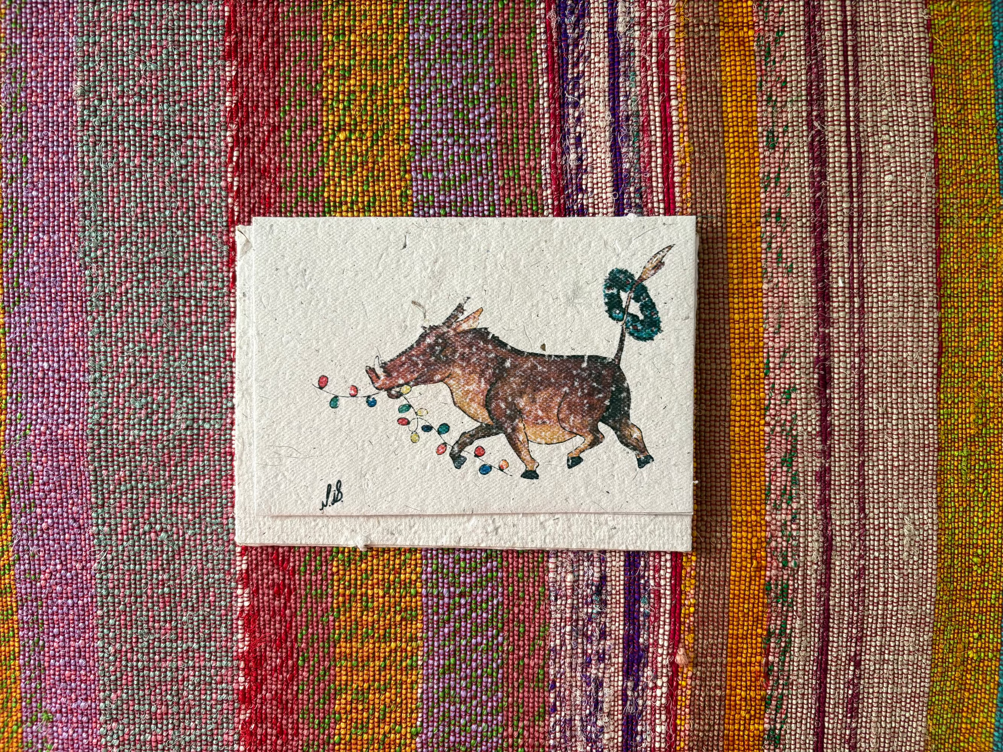 A card featuring a whimsical illustration of a brown cow with colorful dots and a green wreath, placed on a vibrant, multicolored striped woven textile.