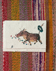 A card featuring a whimsical illustration of a brown cow with colorful dots and a green wreath, placed on a vibrant, multicolored striped woven textile.