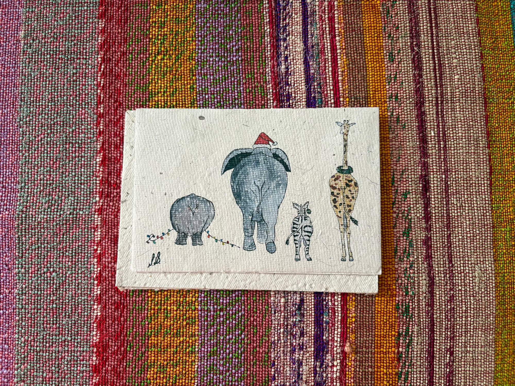 A small handmade paper art print featuring whimsical animal illustrations, including a gray elephant wearing a red Santa hat and a giraffe, displayed on a colorful striped woven textile background.