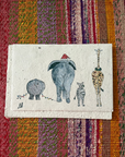 A small handmade paper art print featuring whimsical animal illustrations, including a gray elephant wearing a red Santa hat and a giraffe, displayed on a colorful striped woven textile background.