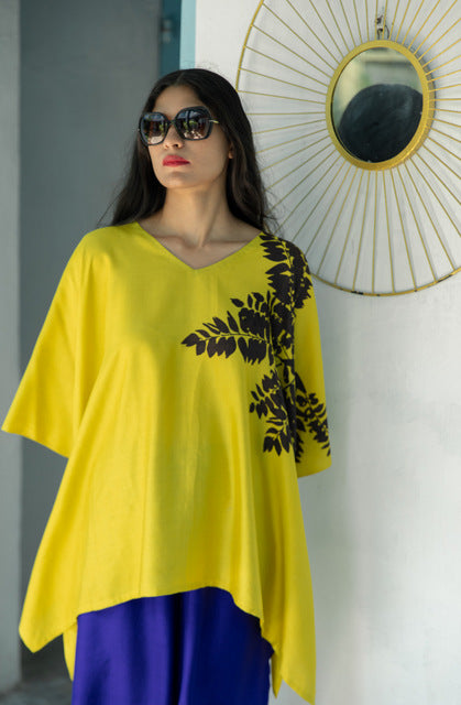 Woman wearing a yellow top with black leaf patterns and blue pants, standing against a white wall with a decorative mirror.