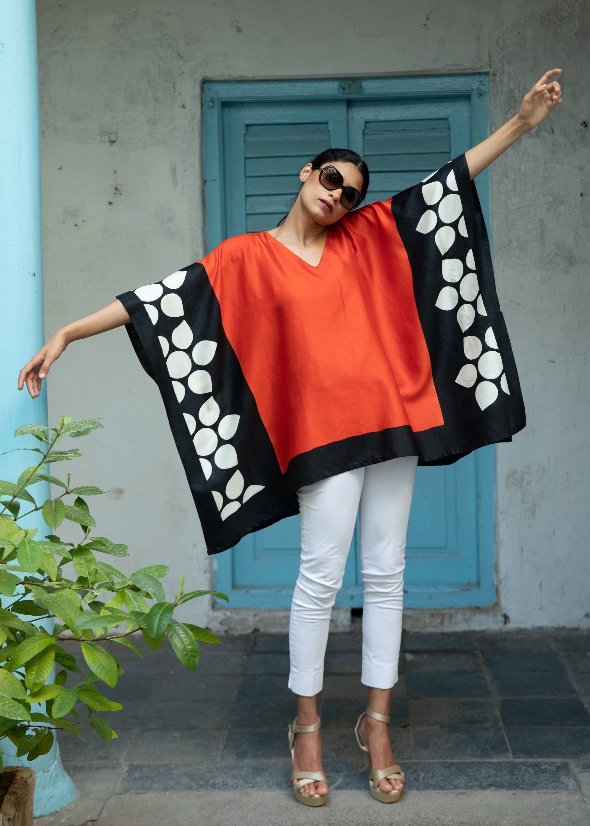 Person wearing a black and orange kaftan with white patterns, standing against a gray wall with a blue door.