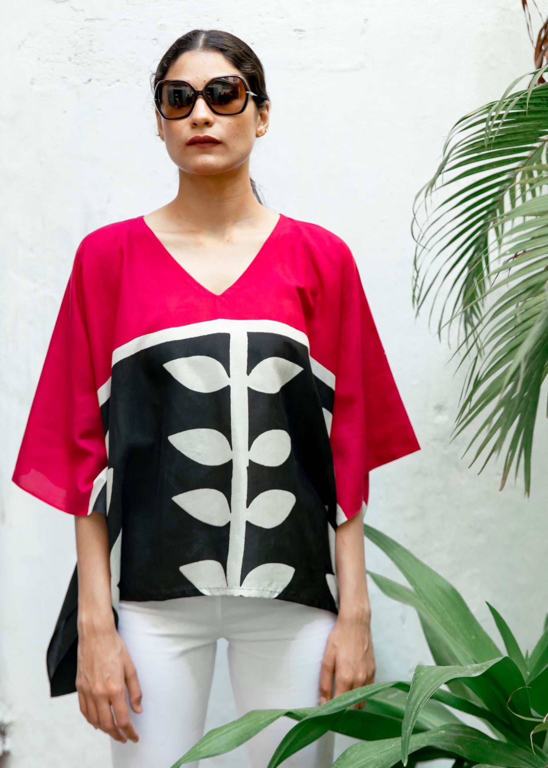 Woman wearing a pink top with black and white leaf pattern, standing against a white wall with plants.