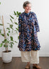 A woman standing next to a potted plant, wearing a navy, aqua, and apricot floral & leaf print dress with button fastening through the front.