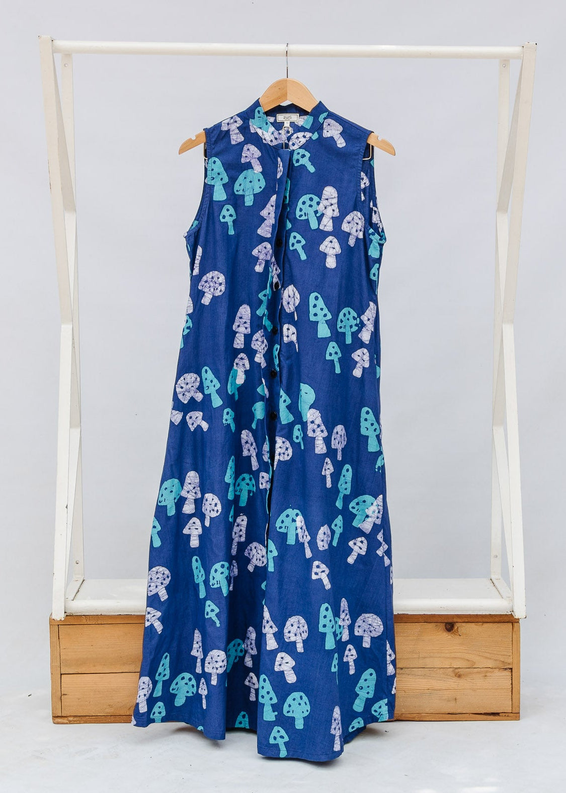 Blue sleeveless dress with white and green patterns on a wooden rack against a white background
