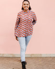 Model wearing blue and red zig zag print long sleeved blouse, paired with jeans and black boots..